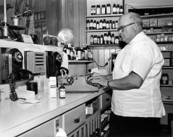 I purchased this 8 x 10 print at the swap meet. On the back is printed:
Mr. Cliff McCorkle, proprietor and pharmacist of the 101 Broadway Pharmacy, Richmond, Calif., preparing a prescription. 5 November 1957. Photographer: Pfc. Barbara A. Warner, Sixth US Army Photo Lab, Presidio of San Francisco, Calif. Official US Army photograph. View full size.
Hey, who ate the cheese?!You have to love the ingenuity of gluing a Victor mousetrap to the drawer and use the "snap" clip to hold what looks to be a hand towel.
PinwormsThe prescription that he is working on (Vanquin) is for pinworms and is supposedly no longer available in the USA. It seems that it caused gastrointestinal problems among other things.
Royal Quiet De Luxe typewriterMy mother had this typewriter from her college days in the mid-fifties. It's rather nice. 
Automatic Electric AE50sNot the jazzy "jukebox" variety with the optional chrome, but just as functional.
[That's about the phones, folks. -tterrace]
Note the fan awhirr -- must have been warm in Richmond that day.
That&#039;s a ZERO fan,which, along with the ESKIMO brand, were mainstay coolers in the Fifties.
Clever names, no?
Popular typewriterThat model, or the 1938 version of it, was used by Jerry Siegel, creator of Superman:
“One of the little known stories about Superman is that he owes a lot of his existence to this typewriter...I liked this typewriter so much that it was the only portable I ever had or used.” -- Jerry Siegel, 1995.
It was also used by Ernest Hemingway and Ian Fleming.
The FanIt's a Zero made by Bersted Mfg. who made several cheapo brands to be sold at drug stores for a few dollars. The quality brands such as GE, Westinghouse and Robbins &amp; Myers were sold in appliance stores.
A Double Take On The PhonesI found one of those phones in an antique store and  installed in our historic (1937) house in Phoenix Arizona about 5 years ago.  It still worked although our grandchildren were clueless about how to use a rotary phone.  You could club someone to death with the Bakelite handset.  It weighed about 2 lbs. 
I have always been fascinatedby how pharmacists were able to get the medicine bottles into the typewriter to create the labels.  Well kept secret at pharmacy school, no doubt.
Zero FanI have a Zero fan that I still use. It rattles a bit, just a little, but it works well. I just have to remember to oil the bearings every summer. 
Built Like a TankWhen I purchased my home in Vancouver, B.C. in 1977, the British Columbia Telephone Company installed this model 50 wall phone in my kitchen. It was originally a rental phone, and when deregulation arrived I purchased this as my home phone for $35.00. While it does not have a chrome dial, it does have the deluxe chrome rings on the heavy handset. This telephone has never needed service in all these years, and it is used a lot. Dial phones came late to Vancouver, and most homes changed from manual exchanges with operators to dial in the 1950s. Originally these phones were without a dial, and as exchanges were converted the "Telephone Man" came 'round to each home and installed a dial. My first phone was in an apartment in 1967, and no private lines were available, so I had a party line for ten years. Such was life with B.C. Tel, a subsidiary of General Telephone at that time. 
No longer a pharmacy, 101 BroadwayLooks like a temp emplyment office now
View Larger Map
Two wall phones?I have a wall phone like those, made by Automatic Electric, the General Telephone equivalent of Western Electric for AT&amp;T. As far as I know, though, Richmond was always Pacific Telephone, an AT&amp;T subsidiary, so why are those phones there? I was born and raised a few blocks from that pharmacy, and am a bit of a phone nut, with a collection of interesting, different phones - like those!
(ShorpyBlog, Member Gallery)