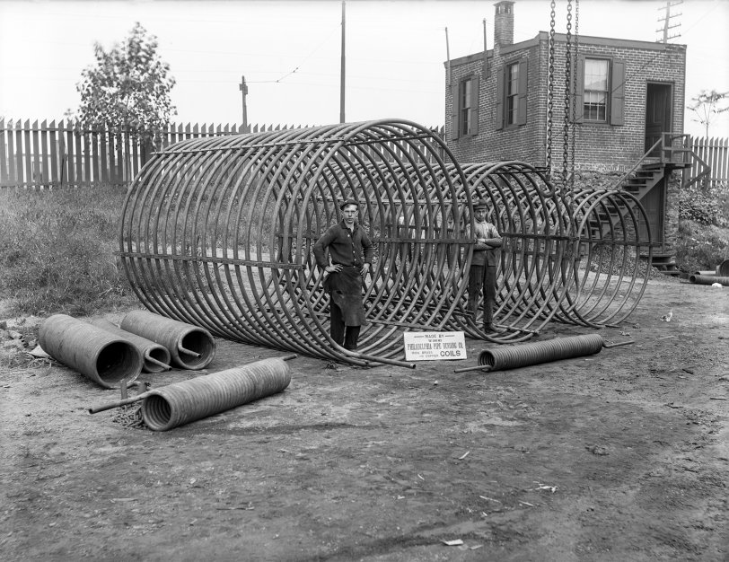 The Philadelphia Pipe Bending Company, founded in 1880, is still in operation today. Here we see two workers standing inside some large coils with advertising signage. 6½ x 8½ inch glass negative. View full size.
