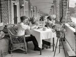 Washington, D.C., 1924. "Chestnut Farms Dairy." ("Cottage cheese, sugar? Yes, honey.") National Photo Company Collection glass negative. View full size.
Are you going to eat that???It looks like she gets her fair share of calories. He, on the other hand, looks like he sticks to the cheese products.
Not a whole lot of lovin&#039;Not a whole lot of lovin' going on with that cottage cheese breath.
Strange CompositionThe photographer seems more fascinated with the perspective of the porches receding to infinity than in the diary-focused dinner.  The odd high table seems to contribute to this. Why not have a normal table height with the camera looking down on the scene?
[It's what happens when you're focusing on that diary. - Dave]
Pshaw on the rowhouse commentThe tunnel-of-doom effect is done with mirrors. It's the same optical illusion anyone sees in a fitting room with mirrors on opposite walls.
You don't see the "people" in the reflections because they're vampires.
Re: Brick TitanicI've often thought my porch and its environs resembled the Lusitania.
Dear DiaryLook again -- the table is normal height.  It's the chairs that are low because they are intended for lounging, not dining.  That is, if you wish to call tomatoes, cottage cheese and milk dining....
Brick TitanicIf it weren't for the fact that it's a dozen years too late and constructed out of material that wouldn't float very well, this could almost pass for the deck of the Titanic.  Not sure if agri-tourism was around back then, but the Chestnut Farms Dairy's estate was positively palatial.  That's a hotel.
[These are rowhouses. The caption note "Chestnut Dairy" means the dairy was the National Photo client who commissioned the picture. - Dave]
Plastic ContainerThe cottage cheese tub looks very similar to those plastic tubs you would find today. Wonder if it is made of tin or paper.
[Waxed paper. - Dave]
Pretty CoolI just got a new cordless mouse and, among other things, it has a magnifier as one of the four possible buttons, plus the wheel. Comes in handy for Shorpy's once in a while. 
Dairy productsYou know, even during Prohibition, I don't think many people outside of dairy advertising drank milk out of wine goblets.
A tidy rowI love the pristine look of these new brick row houses.  This photo reminds me of the 4700 block of 8th Street.

(The Gallery, D.C., Natl Photo)