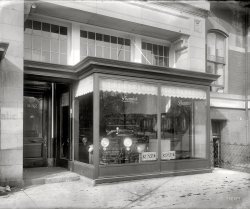 Washington, D.C., circa 1920. "District Motor Co., front." Won't someone give me a home? Or at least a garage. National Photo glass negative. View full size.
I&#039;ll adopt it!I really like the latticed design on the headlight glass covers... I know, that sounds like a weird reason to buy a car; but maybe, if that is indeed a Premier car, and the copy in the attached 1920s ad is true, the fact that "its gears are changed electrically" could be enough to convince the missus that this was not another useless purchase.
CorrespondenceIs that a letter hanging in the mailslot under "932"?
Poor thing, cooped up in therePremier Motor Corporation (of Indianapolis) wasn't in good financial health by July of 1922:

Another ad for Premier, showing the same (or a very similar) model. This ad claims a "magnetic" gear shift.  Sounds sophisticated.
Now concentrateon those amazing and mesmerizing reflector bowls on the head lamps. On the count of three you'll be fast asleep!
A Home?There's a spot in my garage just meant for you!
The Car With Kaleidoscope EyesI can't help but think of the Beatles song, Lucy in the Sky with Diamonds, when I look at those headlights. They are beautiful. It also looks as if someone is seated in the drivers seat, although I realize it is only reflections. Kinda creepy, but still a great shot!
932 14th St., N.W.The location is 932 Fourteenth St., N.W. (streetview).  The storefront, designed by Appleton P. Clark, jr., housed a string of motor vehicle sales rooms.  The building was razed in 1928 to make way for the Ambassador Hotel.



Washington Post, Oct 5, 1913


Will Remodel 2 Buildings
H.K. Willard to Make Improvements to 930 and 932 Fourteenth Street.

Two more of Washington's buildings are to be remodeled and converted into modern business buildings.  The properties at 930 and 932 Fourteenth street northwest are to be entirely reconverted, which will include rebuilding of the front walls and lowering the first floor to the level of the sidewalk.
The windows will also be change in the rear of the first floor, and electricity will be placed in the entire building.  The cost of the work will amount to about $14,000.  The owner of the building is H.K. Willard, and the plans have been drawn by Appleton P. Clarke, jr.  The construction work will be done by Henry Hull.

 The following timeline is culled from adverts in the Washington Post. Killeen's truck sales business had a prior storefront on the same block of 14th street, seen in the Shorpy Post: Worm Drive: 1919.
Apr 20, 1919.
Henderson Motor Car Co.
F.S. Carmody, President.
Franklin 5611.
selling the Columbia Six, Gem of the Highway.
Apr 4, 1920.
W.P. Killeen.
Telephone Frank 6188.
Service Station: 612 L St. N.W.
selling Rainier worm drive trucks.
Oct 21, 1923.
District Motor Company.
Main 620.
Space 13, Fall Show.
selling the new Series U Six-40 Moon.
Advanced TechnologyElectrically shifted gears in 1920, bet that was real reliable. 
The invisible manis leaving footprints!
Rare nowPremier made cars for 30 odd years and apparently there are only about a dozen left. If you want to see a running 1914 Premier see Jay Leno's Big Dog Garage. 
(The Gallery, Cars, Trucks, Buses, D.C., Natl Photo)