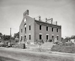 "G.G. Loehler Co., August 1923." The 12th Precinct police station under construction in Washington at 17th Street and Rhode Island Avenue N.E. Cost: $55,376. National Photo Company Collection glass negative. View full size.
MasonryNo prefab walls here.
And here it is today!View Larger Map
RadiatorsOn the left a wood barrel is sitting in a pile of cast iron radiators that would presumably be used in this building for heat. If this building is still standing there is a chance the radiators are still in service. We have similar system still in operation on the campus where I work.
Stacked UpLooks like they delivered the radiators early!
Heavy MetalThe cast iron boiler sections are on the other side of the building.
Free StuffThe most striking difference to me in this photo from any image of a construction site today is that the contractor was able to stack his building materials along the street side of the lot without protecting them with a chain link fence, razor wire, night lighting and guard dogs from nocturnal "salvage" thieves. Radiators, boiler parts, bricks, roof shingles and more are all just sitting there waiting to be hauled off.
My First Thought Was... only the government would spend that kind of money in 1923 on something like that. But it's still there and looking good. Must be one solid structure. I stand corrected.
Thanks for the modern view.Thanks for the modern view. Helps explain why the brickwork was left 'unfinished' under the fascia line. They still had some what we today call 'elevation' applications to put on that area. And it looks like they added on to the back later, with a separate entry. I enjoyed roaming over this building site comparing it to how we do things in construction these days.
The NeighborsI am really surprised to see that the white house to the right in both pictures is still there.  It must have been noisy although they must have felt pretty secure.
First BookingCurious to me that back in the day, a speeding offense resulted in a person being arrested and hauled into the police station.



Teacher First Prisoner At New Police Station

The first man booked at the new Twelfth precinct police station was Marion Richard Vickers, 26 years old, a teacher, living at 1625 K street northwest, who was arrested on Rhode Island avenue yesterday afternoon by Motorcycle Policeman K.L. Potts, charged with speeding.
As Potts came through the door of the new station with his prisoner, Policeman John Donovan, at the desk, assumed a business-like air, and then proceeded to write Vickers' name and address on the first line of the first page of the arrest book.
The new station was opened formally in the morning by Maj. Daniel Sullivan, superintendent of police. In the course of the day many men and women, including several members of the Rhode Island Avenue Citizens association, inspected the station.

Washington Post, Jan 2, 1924 


RadiatorsI have been told that the radiators were usually assembled in place so that only components were moved and not the whole thing. Looks like I've been told wrong.
Additionally, I automatically keep trying to move the original picture around like you do in the Google street view. It doesn't work.
DebarredAppears that the bars on the ground floor windows were removed at some point in the past...apparently it's a safer neighborhood now than it once was.
[The jail bars were to keep the bad guys in, not out. - Dave]
What is thatTapping you guys' knowledge here. The guy on the far right pushing a wheel barrow away from the camera is in front of something stacked up that appears to be sectional square fluted pieces. I cannot figure out what those are. Can any of you I.D. those? Thanks.
[Those are stacked shingles nearest the camera. Behind him it looks like clay conduit, as noted above. - Dave]
Clay ConduitLooks like terra cotta conduit, used to run wires underground.
FireblockThey look like fire blocks; at least that's what we call them. In older commercial buildings, the interior walls were laid up with these block, rather than with wood framimg. A fire-spread preventive device which then got a plaster veneer. Simply hell for rewiring/replumbing projects.
Hollow TileThat's Hollow Tile, kind of a precursor to cinder blocks. Fired out of clay and hollow, they were used in combination with brick to build a load-bearing masonry structure. Grooved so it could accept a plaster finish on the interior face. Fireproofing wasn't the point, although they were certainly fireproof just like brick. Eventually Hollow Tile was supplanted by cinder block construction and brick veneer on a wood stud frame.
(The Gallery, D.C., Natl Photo)