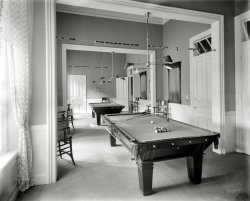 Lake George, New York, circa 1908. "Billiard room, Fort William Henry Hotel." 8x10 inch dry plate glass negative, Detroit Publishing Company. View full size.
Not enough room.A great shot of lovely tables. But this seems to be an afterthought by the hotel. The intrusive column on the right of the pool table would prevent any serious play. Otherwise well equipped, the adaption of a spare corridor was not well thought out.   
AfterthoughtIt looks very tight.  The space doesn't look like it was planned as a Billiard Room from the beginning. Awkward. 
In two delicious flavors!With pockets, or without.
Billiards vs. PoolSome people may not recognize the billiard table in the rear of the photo. It lacks the pockets of its cousin, the pool table. 
Hmmph!Just as I say it takes judgement, brains and maturity to score in a Balke line game, I say that any boob can take and shove a ball in a pocket.
The Mancave.Today I suppose it'd have curtains with a Nascar theme and lights over the tables advertising some beer or another.
Keeping ScoreThe beads on the wire are used to keep score in straight pool or 14.1 continuous pocket billiards, the standard game of the day and the most popular game in America up until about 40 years ago.  My grandfather had the same such counters hung up in his basement over his pool table.  You still see some straight pool played in serious billiard halls, but it has long yielded to nine ball in popularity.
(The Gallery, DPC)