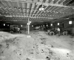 Lake George, New York, circa 1908. "Garage interior, Fort William Henry Hotel." 8x10 inch dry plate glass negative, Detroit Publishing Company. View full size.
BooWhat is that ghostly image on the left? I can almost see a man's face in it. Creepy!
NoticeI can't quite make it out, any chance of a close up?
Marks the spotI've seen a few garage floors in my day, and I don't ever recall seeing more dribbles, drips and spots than are here in 1908.
I'm going to have to look up the date that oil pan gaskets were invented.
The ceilingI can't stop looking at the ceiling! 
If it doesn&#039;t leakthen it's out of oil. My first thought was of the floor of any of my friends' garages and their vintage British sports cars.
I see it tooThe ghostly image on the left might be smoke from an exhaust pipe. Inside it looks like a man that has an open wound in his forehead. Very "Shining" worthy.
Universal DécorI notice the same uninspired 3-lamp lighting accoutrements in the garage as in the Ladies Lounge.
If I saw this in a hotel today, I&#039;d stay elsewhere.While I realize that code required construction techniques have substantially changed building construction, the fact that so many (I count four) of the main support beams are split and failing or (in one case) misaligned and no longer supporting would scare me off this hotel.  And that's discounting the garage itself.
Yes, this style of construction was a firetrap, but this looks to be somewhat less than well constructed beyond that.
[The garage (below) was not part of the hotel. - Dave]
FlivvoplasmThat ghostly manifestation surrounding the spectre of a long-dead Runabout.
Or: a set of piston rings that really need replacing.
Well, I've exhausted that subject.
Drippy carsOil drips were a fact of life with early cars.  Even if gaskets were good most oiling systems were "total loss," meaning what went into the engine or transmission promptly went out of those components right onto the ground. Seals, if any, were a lot more primitive than we have today. If a crankcase got too full you would just drain it, hopefully not on the hotel's garage floor.  The dirt roads absorbed oil drips (and drainings) which helped a little to keep the dust down.
The little red car to the left is a 1908 Model S Ford, the immediate predecessor to the Model T.  How do I know it's red?  That's the color is was offered in.  The transmission was all open and hard to keep lubrication to the working parts.
I don't recognize the other two cars but the near one has some distinctive features.
What Shorpy Will Lead One To !Thanks, Shorpy!
So being from Hartford I decide to track down the Hartford Tires sign hanging in this photo.  After an hour of those typical, wonderful twists and turns I ended up reading about the Hartford Dark Blues, one of the founding teams in the current MLB National League.
Even though I left Hartford for the suburbs at age 5, I was then a bit of a baseball fan (which blossomed with the availability of playing space and teammates in Bloomfield).  Parents and favorite uncle were also big baseball fans (saw my first game at Yankee Stadium at age 5 or 6 and still remember the Yankees beat the Senators by, IIR, 16-5 or so ... the 5 runs in the bottom of the 7th cemented "lucky seventh" in my head forever).
Anyway, I am amazed to just learn that the Dark Blues played at the Hartford Ball Club Grounds which were demolished well before I was born.  But what is astonishing is that site was essentially right across the street from where I lived those first 5 years.  And never was aware of that history.  
Fortunately I do still have crystal clear memories of seeing, outside my bedroom window, that brilliant onion dome on top of the Colt Firearms plant.  I thought that was Turkey or some other exotic land even though it was only about 1500 feet away.
(The Gallery, Cars, Trucks, Buses, DPC)