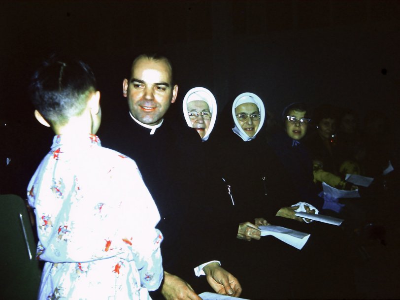 A school concert in 1960 in Penticton, B.C., Canada. Father Quinlin sits with Sister Mary Martin next to him. Beside her is Sister Mary Bridget, the school principal. That is the back of my younger Brother James talking to Father Q. My Dad took the photo.
Sister Mary Martin was my grade 5 teacher. She was older and slow but could hold down our class of 54 kids with just a "look". We always searched that hat outline for just a glance at a hair or two to make sure they were really human and if we ever saw any it was the "buzz" at recess. 
My first meeting with Father Q was when my Mom registered me for grade 1. We went to the rectory and there he was. I was 5 years old and in love. Through the years I always made sure I did not go to his cubical for confession, but if I had no choice I had a rehearsed sin list which was on the light side, because he knew my parents well and I thought he just might tell them something. Although technically that would be a sin, also, and have to be reported to another priest. Somehow that just did not matter to me. View full size.
