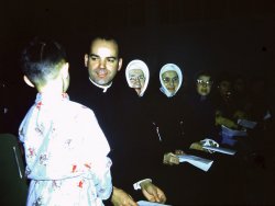 A school concert in 1960 in Penticton, B.C., Canada. Father Quinlin sits with Sister Mary Martin next to him. Beside her is Sister Mary Bridget, the school principal. That is the back of my younger Brother James talking to Father Q. My Dad took the photo.
Sister Mary Martin was my grade 5 teacher. She was older and slow but could hold down our class of 54 kids with just a "look". We always searched that hat outline for just a glance at a hair or two to make sure they were really human and if we ever saw any it was the "buzz" at recess. 
My first meeting with Father Q was when my Mom registered me for grade 1. We went to the rectory and there he was. I was 5 years old and in love. Through the years I always made sure I did not go to his cubical for confession, but if I had no choice I had a rehearsed sin list which was on the light side, because he knew my parents well and I thought he just might tell them something. Although technically that would be a sin, also, and have to be reported to another priest. Somehow that just did not matter to me. View full size.
(ShorpyBlog, Member Gallery)