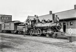 Circa 1899. "Tracy, Minnesota -- engine of the South Dakota division, Chicago &amp; North-Western R.R." Detroit Publishing glass negative. View full size.
Number 605Looks so new it must still have the price sticker on the window. Magnificient detail.
Pressed Paper?Solidly in the link and pin era, the front shank coupler announces this is pre-1900 when such couplers were finally banned.Note the "door" hinged to the side of the headlight. When waiting in a siding, the fireboy went out on the pilot to close the door on the lit headlight so as not to blind the approaching train. This was actually easier than turning out the oil or acetylene lamp. I've seen roller shades used here most often; I can't recall seeing a hinged door before...eh, but those CNW guys could be funny.
Note the headlight mounts are left over from the short smokebox of the engine's original arrangement.Directly below the headlight on the boiler front are two brackets; these were most likely used to brace a pilot-mounted snowplow, necessary equipment in the Plains.
You can almost read the builder's plate date and shop number, but she was built by Schenectady Locomotive Works. Lastly, the front wheels are made of...get this...pressed paper.Look it up, I ain't fibbing![pressed paper railroad wheel]
Schenectady Locomotive Worksthat manufactured C&amp;NW engine #605 operated from 1848 until it merged into American Locomotive Company in 1901.  One of the engines it manufactured for C&amp;NW (#1015 in 1900) is still in existence at the Museum of Transportion in Kirkwood, MO.
Actually, locos were kept this clean &#039;back in the day&#039;>Looks so new it must still have the price sticker on the window. 
Although it's possible this was recently out-shopped, it's much more likely it came fresh from the roundhouse.  Back in the 19th century, locomotives were kept in pristine condition; railroads hired people to wipe them down and engineers took particular pride in the appearance of their engines.  
Olde Buck points out this was most likely converted from wood to coal, I'd guess this loco was constructed in the early 1880s and converted in the late 1880s/1890s.
Note the fixture for a steam line on the roof of the baggage/mail car and the corresponding rig on the back of the tender.  There's a steam feed from the steam dome (alongside the whistle).  I think this was probably added to the original locomotive.
There's a 'mail bag catcher' on the door for mailbags.  I think we've had previous Shorpy photos showing the mail bag hangers alongside a passenger depot.
Magnificent Locomotive!And it's obviously being maintained by a crew that really cares about it.  As Olde Buck implies, it is probably not new -- in 1899, the 4-4-0 layout was already considered classic if not obsolescent, and the pin connectors, notoriously dangerous, were being replaced by the "modern" type of couplers. Note the brightly polished bell, the shining surface of the boiler, and other authentic details -- you can see the whistle on top of the steam dome and the lever that triggers it with a lanyard leading back to the cab.  The sand dome shows the plumbing that deposits sand on the fore driver wheel as needed for traction.
I don't know how to recognize it in railroad wheels, but it is certainly true pressed paper was used in some, with an iron "tire" on the rim.  In the 1880's, pressed paper was even used for the skins of racing shells, as it was lighter than wood.  I don't think it is used today for any structural purposes.
In case of fireCheck out the fire escape on the side of the hotel in the background.It doesn't look too fireproof itself! 
Not PaperThe pressed paper hubs were strictly used on Pullman Sleepers after
the inventor showed George that they really did reduce vibration transmission.
I could be wrong butI bet it has that new locomotive smell.
During World War Two plastic-reinforced jettisonable paper fuel tanks were used to extend the range of fighter aircraft including the P-47, P-51 and P-38. The largest held 200 gallons, which gave (for example) the P-47 close to a couple more hours of flying time, enough to escort our bombers over Germany. This ability was a very significant contributor to winning the air war because enemy fighters no longer could easily attack the big and unprotected B-17s and B-24s, unprotected except for their many .50 caliber machine guns (some B-17s packed 11 or more), which made many enemy pilots moribund.  
Pressed paper wheels> I don't know how to recognize it in railroad wheels, but it is certainly true pressed paper was used in some, with an iron "tire" on the rim.
The give-away is the bolts/rivets on the leading truck wheels.  These are typical of composite wheels.  A cast wheel would not need these bolts/rivets to hold it together.  
Builder&#039;s engine  DataC&amp;NW 605 was built by Schenectady Locomotive Works in 1885. C&amp;NW labelled it  C-2 class. Boiler pressure was 150 lbs.  Driving wheels were 63 inches diameter.  605 was scrapped March 5, 1917. 605 weighed 88,500 pounds.  Tractive effort was 16,900 lbs. Looks from my source that the railroad had only a dozen C-2 class engines.  
A Work In ProgressWhoever was doing the sprucing up must not have had time to finish the tender before the engine was needed for service, because the old, dull finish is visible around and inside its numbers, and on the curved gangway area where the crew climbs up to board.
JugI wonder what beverage was contained in the earthenware jug aft of the engineer.  
Second career for a C&amp;NW C-2 4-4-0 - can you help?At least one of these C&amp;NW C-2 class locomotives was sold secondhand.  I have a large B&amp;W print of Sheffield &amp; Tionesta #4 at Kelletville, PA in 1904, with no visible builders plate, but "C&amp;NW" cast in the smokebox door just as in 605.   S&amp;T #4 was used between 1904 and 1914 as the primary passenger engine on the little S&amp;T, and was replaced by a new Baldwin 4-4-0.  The entire railroad was abandoned and sold for scrap in 1941.
Judging by the matching set of dents on the air compressor on the photo I have taken from the same angle, I seriously thought S&amp;T 4 was C&amp;NW 605.  If there's any information anyone can find on which one it was, contact me.
Sheffield &amp; Tionesta is convered in the book "Teddy Collins Empire" (Casler, 1974) of the "Logging Railroads of Pennsylvania" series.  His book is out of print but the railroad museum of PA has the negatives and original manuscript.
Finding this particular image on Shorpy was wonderful and made me a member.
(The Gallery, DPC, Railroads)