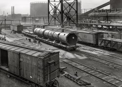 April 1943. "Detroit, Michigan. A huge pipe section loaded on a flatcar." 4x5 inch acetate negative by Arthur Siegel for the Office of War Information. View full size.
Plus our old friend the gasometerAs seen previously on Shorpy - Gasometers
Standing GuardThe soldier seems fascinated by the man doing something to the rails. I wonder what branch of the Army the soldier is? And why is he standing guard? 
Don&#039;t try anything funny with that torch!Under the watchful eye of the soldier on the left. This scene is at the 'RIP' track (Repair In Place) so perhaps some of the blocking or load tie downs needed attention on the precious cargo on the flat car. Since the load is longer than the car it is mounted to an 'idler' car is needed at the far end which also provided added space for additional lading.
Flatcar loadPipe, schmipe, I want to know what the load on the following flatcar is. Looks like the backbone of a giant monster- perhaps a new secret weapon that explains the soldier and his rifle off to the left.
It&#039;s a column, not a pipeIt looks like a distillation column. The flared section at the near end of the flatcar is the skirt that will support the column when vertically installed on its pad. You can see a series of flanged necks on the sides of the straight sections that either correspond to internal plates in the column when components of the feed mixture are removed or where feedstocks are added for refining. 
Saturn V section?And the state of the art 1940s refrigeration. The ice car's right behind the booster ...
&quot;Pipe, Schmipe&quot;, IndeedI concur "hugely" with OVBuckeye that we see a brand-new distillation column on its way to who-knows-where -- any of a number of Great Lakes region petroleum refineries would be my guess.  I've worked in the fine chemical industry for decades and I know a distillation column when I see one.  In fact, I think there are some smaller diameter columns on the next car beyond.
(The Gallery, Arthur Siegel, Detroit Photos, Railroads)