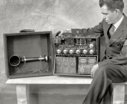 Washington, D.C., circa 1924. "Brent Daniel, formerly of the Radio Laboratory of the Bureau of Standards at Washington, with the first portable Super-Heterodyne, his own design. The seven vacuum tubes, batteries, loop antenna, loudspeaker and other necessary units are completely self-contained in the carrying case. He has been able to hear Pacific Coast stations from this outfit." View full size.
BatteriesThe batteries on the right power the low voltage high current filaments, probably 6v in parallel; the batteries on the left supply probably 48v each in series for a jolting 144 volts for plate current.  Keep your fingers out.
Batteries were the problem; they're heavy, expensive and costly to replace, which they are often.
If you can't pick up anything, you can at least keep yourself warm from the heat it throws off.
[The 15-cell batteries on the left are 22.5 volts each, or 1.5V per cell. "Heavy Duty 6" on the right are labeled 1.5 volts (per cell, I guess). - Dave]
The ultimate in portabilityIt's the size of a Breadbox and only weighs fifty pounds!
You just know he was thinking that they'll never get any smaller than this!
The latest technology..."Transistors Under Glass"
Portable, all rightIf you have a dolly!
And thirty years later.It came down to this size. This is the first commercial transistor radio sold in 1954 by Regency. I remember listening to many a Giants game one those transistors. I wasn't around for the one in the main photo.
ReflectionsI love it when one of these old photos contains a reflective surface that provides a bit of unintentional insight into the background.  In this case, each of the silvery tubes tells a slightly different story - depending on which one you look at, you can see the subject's hands, legs, and feet, the camera and a bit of the photographer, the large window that runs the length of the studio, some sort of lamp that's projecting a halo of light onto the ceiling, various bits of furniture and shelving, and if I'm not mistaken, part of the (adjoining?) building outside the window.
At least we can figure it out!Unlike an iPod, at least the components are somewhat easy to understand. Wonder what he would think of an iPod?
Complete in Itself


Washington Post, July 15, 1923.

Make Successful Test of
Portable Suitcase Radio Set


Local Enthusiasts Get Clear Reception
 With Type Built by Brent Daniel


Various types of portable radio receiving sets have appeared from time to time in the last few months. While taking different forms, all the sets require either an external coil aerial or overhead antenna when in use, thus limiting their use to stationary installations or specially equipped conveyances.

A Washington manufacturer recently has standardized a design of portable receiving set which is complete in itself. The entire outfit, including all the batteries and coil aerial, is contained in a medium-sized light-weight suitcase.

This portable receiver is ready for important use at any time by merely closing the switch which lights the filaments of the six UV199 vacuum tubes used in the radio audio amplifier receiver. This amplifier employs three stages of DX-12 radio frequency transformers, detector and two audio stages. The same type instrument in nonportable form has been used repeatedly in the reception of transcontinental radiophone signals by employing a three-foot square coil aerial.

The builder of this portable set, Brent Daniel, recently made a series of tests to determine the practicability of its use in an automobile in motion, and in general outdoor reception. WCAP, the Chesapeake and Potomac Telephone station, was tuned in when the set was located on the fourth floor of an office building. Leaving the set in operation with the musical program from the broadcasting station coming in clear and loud, the set was carried downstairs and placed in an automobile without once interrupting the reception. When the car started off, the ignition spark was quite audible with the set placed near the bed of the machine, however, by placing it in different positions, the spark was not audible. &hellip; 

These tests, and the reception accomplished later with this portable set demonstrated its scope of usefulness in the hands of the auto-tourist or vacationist. With a high-class broadcasting station within a few hundred miles range, the user of such a set is assured of entertainment, regardless of his location. &hellip; 

The outstanding feature of this portable receiver is that when the summer season is over and a radio set is wanted for the home during the winter, it is only necessary to remove the amplifier receiver unit from the portable case and place it in the regular mahogany case which is furnished for indoor use.
 
The superheterodyne receiverThe superheterodyne receiver and FM radio were both invented by Edwin Armstrong, generally acknowledged the greatest American radio engineer. A Signal Corps officer in WWI, he gave his patents to the US government during both WWI and WWII. FM radio contributed to Allied victory in WWII since it provided communications when AM did not. Armstrong lost a years-long patent fight with RCA and committed suicide in 1954. His wife continued the fight and eventually won the FM patents from RCA. Armstrong's life is documented in the book and Ken Burns' film "Empire of the Air". 
SuperheterodyneThe big word "superheterodyne" means that this radio was quite advanced over the common tuned radio frequency (TRF) radio of the day. The fact that it has seven tubes and only two tuning knobs is unique among 1920s radios. Later, the two tuning knobs were put on a common shaft, allowing the single knob we're used to. 
The trick used in this model is that it converts the station's frequency to a lower intermediate frequency that's the same no matter which station you're listening to, and amplifies the weak signal using an amplifier that's factory-tuned to that one frequency, instead of requiring the listener to tune several amplifier stages to the station's frequency. Hence the need for fewer tuning knobs. 
By 1935, after the patent mess got sorted out, this type of radio completely replaced the TRF radio that Atwater Kent made a fortune from. It later became known as the All-American five tube radio, after the bean counters whittled every last penny from the design in the late thirties.
Ultimate geekHe's even got his pocket pen protector!  This must have been very advanced for 1924.
Dry Cell BatteriesZinc-carbon chemistry gives 1.5 volts per cell, so all batteries made up from them in series will be multiples of that.  The "A" battery was for the filaments, "B" for the high plate voltage (often 90V), and "C" for the grid circuits of the vacuum tubes (often 67.5V, so three of those 22.5 bats in this - four would give the 90V).  The UV-199s (triodes) in it have 3V filaments, drawing 0.06 amp each.  Larger current capacity is achieved by using cell electrodes with more surface area (so larger and heavier cells) or by connecting smaller cells/bats in parallel.  On those large "Twin Cells" on the right their terminals are marked "carbon +" and "zinc -".  Burgess has an interesting history with its distinctive "zebra striped" product.
Zenith TransoceanicThe largest radio I sold was the Zenith Transoceanic Radio. Zenith produced this shortwave radio that could be powered by AC or batteries . It was first produced in 1942 and continued to be made until 1981. In 1960 it sold with the batteries for about $160, figuring the average U.S. wage at the time at about $4000, it cost about 2 weeks pay. It weighed in excess of 25 lbs.
Not the only portableI happen to be the owner of a RCA Radiola AR-812, which is considered the first commercially produced superhet, and, which was considered a portable radio. Production began in 1924, and the radio sold for the bargain price of $220! It's an enormous thing, one foot high by one foot deep by three feet wide, with a mahogany veneer. I have a wonderful ad somewhere showing two guys camping, with fish in the pan, "listening to the big game". The tubes are encased in beeswax to protect them during transport. This feller's is a homebrew model. To understand the importance of the superhetrodyne technology, one must realize that there were several competing systems vying for dominance when the superhet came out. Today, if you have a radio, it's a superhet, period. 
Many of the facets of radio we take as given today were not yet common when these radios were being built. For example, the mHz system was not yet adapted, so every single knob on the radio scales from 0-100. Volume knobs, tuning knobs, everything. Gotta love it.
[Spelling note: heterodyne, not "hetrodyne." - Dave]
(Technology, The Gallery, D.C., Harris + Ewing)