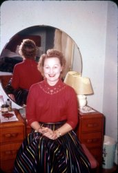 1941 - Arlene "Billie" (Wogatske) Morrell. 5 Bellaire Dr, Old Saybrook, CT. 35mm Kodachrome slide. View full size.
Time capsuleThis photo is chock-full of awesomeness.  The art deco vanity, frilly curtains seen in the mirror, doilies carefully positioned on the vanity, grandma's shiny ribbon skirt and gold-trimmed blouse, her bright red lipstick and big smile...it's all fantastic. It looks like a fur coat is thrown across the bed, so maybe she's getting ready for a night out on the town.  I like the way the photographer carefully positioned himself/herself just out of range of the mirror, too.
DecorLove, love, love that vanity. And I remember those decorative tin wastebaskets from my grandfather's house. Nowadays, trash cans are plastic and boring. Great pic! 
(ShorpyBlog, Member Gallery)