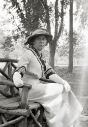 From the collection of film and 4x5 glass negatives I recently purchased that seem to have been taken in upstate New York about 1912. This is from a film negative that has been bent, hence the flare on the right side.  The lady's dress is spectacular, and she is coordinated with gloves and white shoes.  I don't think this was an inexpensive outfit. View full size.
Sunday Best Maybe?Could this be a Sunday best outfit? Maybe visiting the in-laws? Either way, beautiful. The camera definitely loves this lady, bend or no bend in the negative.
FabricMy guess would be that the dress is linen, which was a common fabric for "day dresses" like this one. Linen is pretty comfortable/breathable in hot weather, even in a fairly heavy weave. It's also very prone to wrinkling, so this lady's perfectly pressed dress may also be well-starched.
Smile, dammit, smileI know nobody cares what I think and I know many people who would never allow themselves to crack a smile in their posed pictures because it may not be "cool" or sophisticated, but this elegant lady would look a million times better, younger and healthier if only she would have smiled or at least put on a pleasant face.  Instead she has a dour expression in both of the photos we've seen that makes her look gloomy and judgmental, with a superior attitude.  Even Mona Lisa looks more approachable. Perhaps we will see a happy picture of her in the future. (That's my story and I'm stickin' to it).
[Try this one. -tterrace]
Colors?Seeing this vision of sartorial splendor, I wonder what she looked like in color. Gloves are obviously white; dress is off white, probably a light cream color. The cuffs and collar make the dress. I'm thinking gold braid over a deep midnight blue. Stunning indeed.
Was this same lady...... in the rocking chair a few photos earlier?
[Yes. - tterrace]
Lady in RedNatural beauty.  Does not appear to be wearing any makeup or lipstick.   For color, I'm guessing dark red or maroon cuffs with gold braid.  Hat is red, dress is off white.  
A Lady, IndeedCarriage, bearing, clothing...everything about her says she's had the best all her life, including education. And in 8 more years she'll be able to vote. How much talent in America was wasted while living in men's shadows?
Yes, but what is the fabricWhat is the fabric the dress is made from? Very thick and stiff but pliable - and warm?
A ladywith style and determination, judging by the look on her face.
Saranac Lake?The background looks a bit too bucolic for Saratoga. And the bench looks a bit rough-hewn for Chautauqua. Saranac Lake was a summer destination for wealthy New Yorkers; possibly this series was taken there.
Mohonk Mountain House?When I saw the first photo of the lady sitting in the rocking chair, it sparked a thought in my mind but I let it pass for the moment.
Now seeing the second photo of the lady sitting on a rough-hewn bench, I am even more suspicious.
There is a resort in New Paltz, NY called the Mohionk Mountain House. Like me, anyone who has ever visited there is familiar with all the rocking chairs on the porches, and the rough-hewn benches and gazebos along the walking paths.
I wonder if this is where these photos were taken?
Going to the website for the hotel will produce an abundance of photos with the rocking chairs and benches I have alluded to....
Dress commentsMy wife and I have done historical costuming for years. The fabric of this dress is almost certainly worsted wool. It's surface smoothness looks like worsted and the folds and drapes are too full to be even heavy linen. The dress she's wearing in the first photo posted (the eyelet lace dress) is either linen or cotton or possibly even a blend of the two.
As to colors, that's harder to determine than you might think. Early black and white films didn't respond to colors in quite the way we expect nowadays. Silver halide is most responsive to the blue end of the spectrum. Colors from the other end often wound up overly dark. With glass plate negatives, special dies and filters could be used to produce a more accurate tonal map of the colors, but those didn't work with film at this time. 
It wouldn't surprise me if the dress was actually bright blue with bright yellow collar and cuffs or something like that. Wool takes die especially well and so was often very brightly colored (particularly after the invention of aniline dyes in the 1850s).
(ShorpyBlog, Member Gallery)